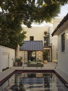 A house with a white exterior and a backyard featuring a pool with a blue tiled bottom, surrounded by a patio with a couch and chair, and a dining table and potted plant