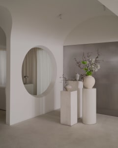 A minimalist living space with white walls, a large mirror, and a vase with flowers on a pedestal, set against a white floor