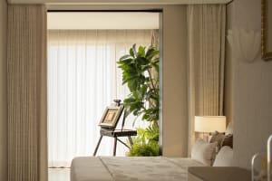 A well-lit bedroom with a bed, a potted plant, and a painting on a stand, surrounded by white curtains and wood accents