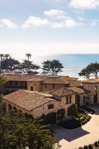A large Spanish-style house with a stone and brick exterior, surrounded by a beach setting with the ocean visible in the background, with natural light and a warm atmosphere
