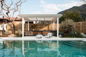 A serene pool area with a pool house, surrounded by a fence and potted plants, featuring a bench, chair, and dining table