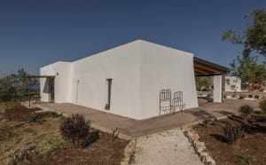Exterior view of a modern minimalist white stucco villa with a covered terrace, black metal chairs, and surrounding olive trees on a hillside.