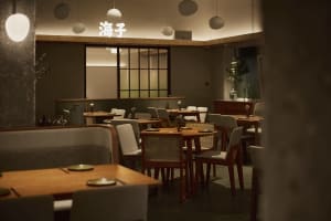 Interior view of a contemporary Japanese restaurant featuring wooden tables, upholstered and cane-back chairs, concrete pillars, and warm pendant lighting with Chinese characters illuminated on the back wall.