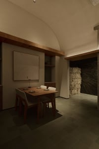 Modern wabi-sabi dining room featuring a wooden dining table with four upholstered chairs, a large topographic wood artwork on the wall, built-in wooden shelving, and a striking wall of hay bales under a high curved concrete ceiling with warm ambient lighting.