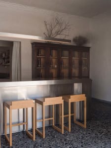 Modern bar area featuring three light oak stools in front of a stainless steel counter, with a large dark wood glass display cabinet in the background, dried branches as decor, and dark terrazzo flooring.