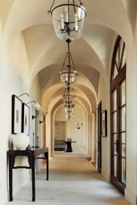 Long arched hallway in Tuscan style featuring multiple glass and iron lanterns, a distressed black console table with white ceramic vase, framed artwork, and limestone flooring leading to a seating area.