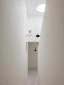 View looking up into a minimalist white double-height hallway featuring two black spherical pendant lights, a classical bust on a high ledge, and a large circular skylight.