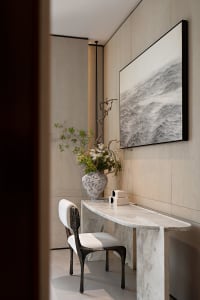 Minimalist entryway featuring a curved white marble console table, a dark sculptural chair, a large ceramic vase with floral arrangement, and a black-framed ink wash painting of waves on a light wood-paneled wall.