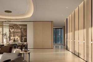 Modern open-plan dining and living space featuring a large circular chandelier, neutral upholstered seating, a round dining table, and a prominent black-and-white mountain ink painting on a vertically ribbed wall, complemented by warm oak paneling and a glossy light floor.