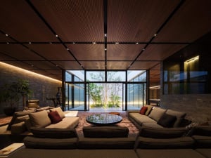 Modern luxury living room featuring dark wood slat ceiling, large sectional sofas, round coffee table, and floor-to-ceiling glass doors opening to a private garden with trees.