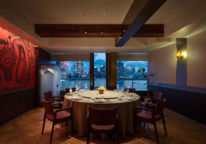 Elegant private dining room featuring a large round table with red leather chairs, dramatic red marble wall, dark wood beams, and large windows showing a twilight harbor city view.