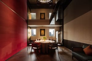 Modern private dining room featuring a bold red lacquer wall, round dining table with six chairs, double-height ceiling, and dark wood accents.
