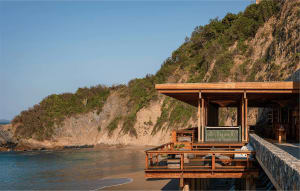 Modern teak wood deck and pavilion built over the ocean at the base of a rocky cliff with lush vegetation under a clear blue sky.