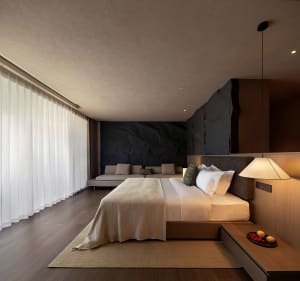 Modern minimalist bedroom with large bed dressed in white and beige linens, dramatic black natural stone feature wall, built-in wooden nightstand, pendant lamp, and sheer white curtains along a full-height window.