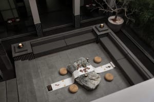 Modern minimalist outdoor courtyard with grey stone pavers, built-in grey benches, large natural rocks, woven straw cushions, and a white embroidered runner set up for a Japanese tea ceremony at night.