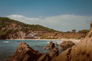 Modern concrete modular resort buildings integrated into a green hillside above a sandy beach and turquoise ocean, viewed from rocky outcrops in the foreground.