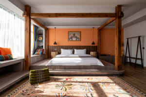 Bright bohemian bedroom featuring a large wooden four-poster bed, burnt orange accent wall, white linens, and colorful patterned rug with sunlight casting strong shadows.