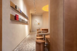 Narrow minimalist cafe interior with terrazzo tile floor, wooden bar stools, brass floating shelves displaying books, and warm ambient lighting.