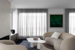 Contemporary living room with beige boucle curved sofa, white irregular marble coffee table, dark blue rug, sheer curtains, and large green abstract painting.