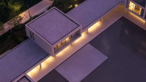 Aerial night view of a modern minimalist concrete villa with flat gravel roof, dramatic linear LED lighting along the eaves, and a large reflecting pool.