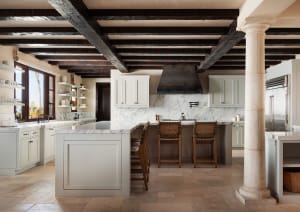 Bright and airy kitchen interior with a large white marble island, three woven leather bar stools, white shaker cabinets, marble backsplash, dark reclaimed wood ceiling beams, and a stone column.