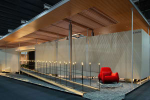 Modern exhibition booth featuring a vibrant red velvet armchair, warm wooden slat ceiling, textured beige walls, and dramatic uplighting along a raised walkway.
