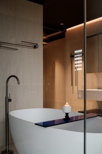 Modern minimalist bathroom featuring a white freestanding oval bathtub, tall black floor-mounted faucet, travertine stone walls, and a lit candle on the tub edge.
