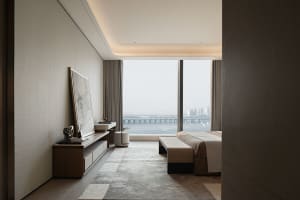 Modern luxury bedroom interior featuring a large harbor view window, walnut media console, textured area rug, and woven bench at the foot of the bed.