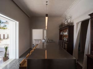Long stainless steel kitchen island used as a home bar in a bright, minimalist interior with pendant lighting and dark wood cabinetry