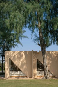 Modern minimalist building with beige sandstone walls featuring large angular triangular windows revealing white sofas inside, framed by a tall pine tree against a clear blue sky.