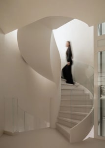 Modern interior featuring a sculptural white spiral staircase with smooth plaster curves and glass railing, a woman walking upstairs.