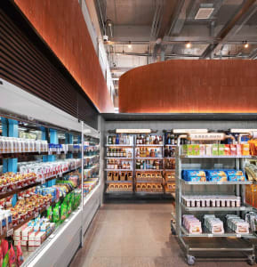 Modern convenience store interior with a large curved Corten steel wall, refrigerated display cases on the left, and industrial metal shelving units displaying liquor, snacks, and daily goods.
