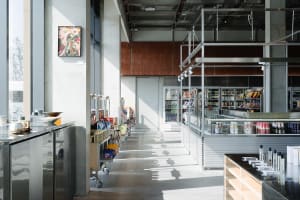 Modern industrial-style convenience store interior with polished concrete floors, stainless steel counters, open shelving, and a second-floor mezzanine structure.