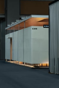 Modern minimalist exhibition booth made of large light-gray concrete panels with integrated warm LED lighting running along the base and wooden ceiling detail.