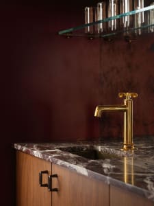 Close-up of a luxury bathroom vanity with deep burgundy marble countertop and integrated sink, paired with a polished brass faucet against a matching burgundy wall and glass shelf above.