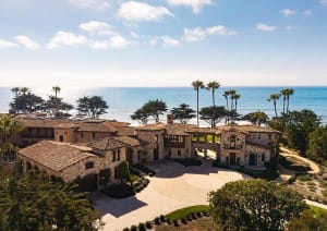 Aerial view of a luxurious Mediterranean-style stone villa with terracotta tile roofs, circular driveway, and lush landscaping overlooking the blue Pacific Ocean under a clear sky.