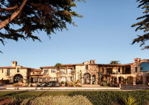 Front view of a luxurious Tuscan-style stone villa with multiple arches, terracotta roof tiles, and landscaped courtyard under a clear blue sky.