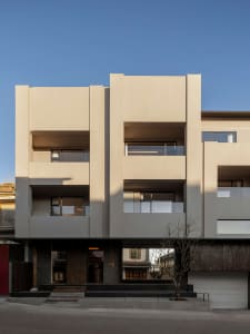 Front elevation of a modern minimalist concrete residential building with recessed balconies and dark wood accents under a clear blue sky.