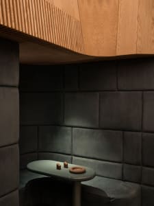 Intimate curved seating nook featuring dark charcoal velvet padded walls, a light oak fluted ceiling, and a sage green oval table with decorative objects.