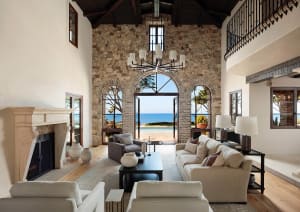 Spacious coastal living room with high vaulted wooden ceiling, textured limestone walls, large beige sectional sofa, modern chandelier, and panoramic ocean view through arched glass doors.