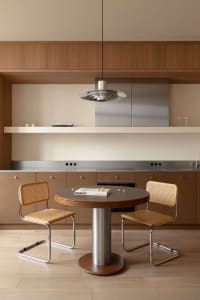 Modern minimalist dining area with a round walnut table, two cane and chrome chairs, stainless steel kitchen counter, and wooden upper cabinets under a sculptural metallic pendant lamp.