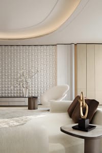 Modern luxury living room featuring a dramatic white 3D textured geometric wall, curved white lounge chair, cream sectional sofa with brown velvet pillows, and a sculptural gold knot ornament.