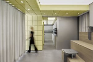 Modern shoe store interior with lime green perforated acoustic wall panels, white curtains, wooden bench, grey stool, and recessed ceiling lighting.
