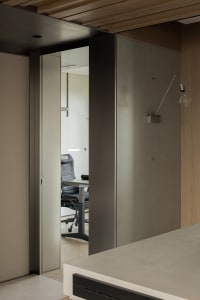 Modern minimalist home office viewed through an open sliding door, featuring neutral wood paneling, a black ergonomic office chair, and a wall-mounted swing-arm lamp.