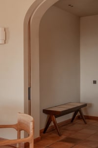 A serene entryway featuring a dark walnut bench with woven cane seat, terracotta floor tiles, arched doorway with wood trim, and a wooden chair in the foreground.