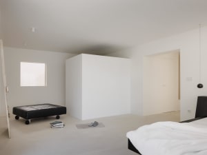 Minimalist bedroom interior with white walls, polished concrete floor, black low platform bed with white bedding, black leather rolling ottoman, and a small square window providing natural light.