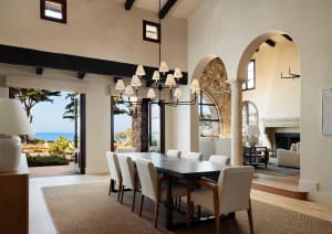 Spacious coastal Mediterranean dining room featuring a long dark wood table, eight upholstered chairs, exposed black ceiling beams, large arched openings revealing ocean view, and a stone fireplace in the background.