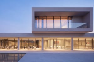 Exterior view of a modern two-story concrete luxury villa featuring full-height glass walls, a second-floor cantilevered balcony with glass railing, and a reflecting pool in the foreground at dusk.