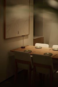 Modern minimalist dining room featuring a solid oak table, four upholstered chairs with wooden frames, a sculptural table lamp, and a large framed Japanese line-drawing wall art with calligraphy.
