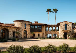 Front view of a luxurious Tuscan-style stone villa featuring arched entrances, wooden balconies, terracotta roof tiles, and a spacious cobblestone courtyard lined with palm trees under a clear blue sky.